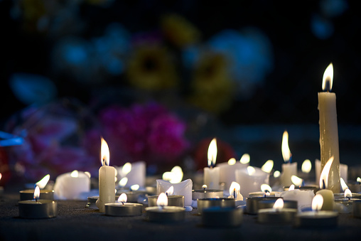 A growing makeshift vigil for victims made out of various size candles on a concrete sidewalk at night with flowers and some flowers stuck in the chainlink fence in the background. These types of memorial vigils happen after a tragic event such as a mass shootings, terrorists acts or accidents.