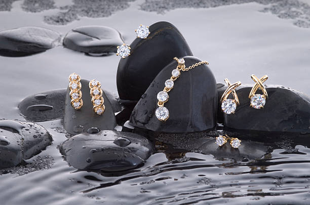 Making a Splash With Gold and Diamonds An assortment of gold diamond earrings with an eternity necklace laying upon a bed of black rocks and rippled water. gold jewelry stock pictures, royalty-free photos & images