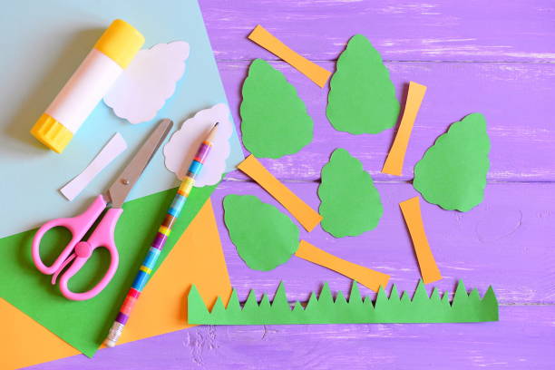 Making green Earth day card. Step. Tutorial. Earth day trees and grass cut from colored paper, scissors, glue stick, pencil, template, paper sheets on a wooden table background. Earth day paper crafts concept for kids. Top view stock photo