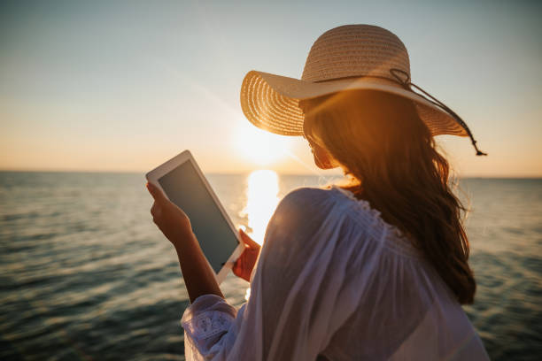 Making memories Beautiful young woman in white sun dress and with hat taking selfie with her digital tablet on the beach at beautiful summer sunset above the sea summer girl stock pictures, royalty-free photos & images