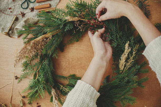 Making rustic Christmas wreath flat lay. Hands holding fir branches, and pine cones, thread, berries, scissors on wooden table. Christmas wreath workshop. Authentic stylish still life stock photo
