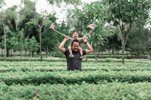 Malay boy holding Malaysia flags carried by his father on the shoulder enjoying morning sunlight at the park stock photo
