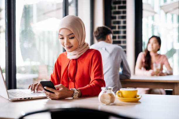 Malaysian woman in cafe using electronic banking on laptop Malaysian woman with hijab sitting in cafe business Malaysia stock pictures, royalty-free photos & images