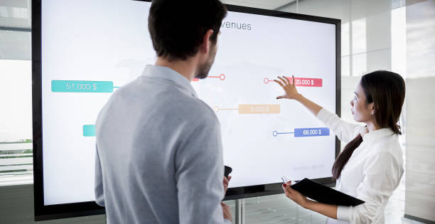 Male and female colleague discussing financial diagrams on large screen in meeting room and preparing for presentation Male and female colleague standing by large screen in meeting room, going over financial presentation shown on screen in meeting room. touch screen stock pictures, royalty-free photos & images