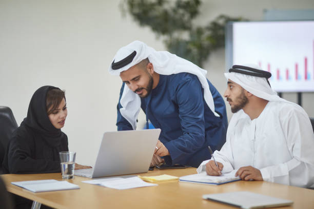 Male and female colleagues discussing over laptop Businesswoman using laptop while discussing with colleagues at desk. Male and female professionals are planning at desk. They are in office. abaya clothing stock pictures, royalty-free photos & images