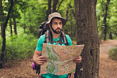 Male brunet bearded confused tourist got lost in the forest, holding map, looking far, trying to find the way. He has a backpack, all needed for overnight stay. Search the trail way