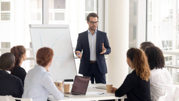 Male business coach speaker in suit give flipchart presentation Male business coach speaker in suit give flipchart presentation, speaker presenter consulting training persuading employees client group, mentor leader explain graph strategy at team meeting workshop Leadership stock pictures, royalty-free photos & images