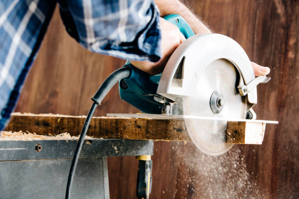 Male carpenter using electric circular saw in home workshop with wood chips flying stock photo