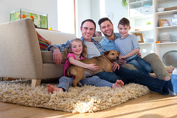 Male couple posing with son, daughter and dog Same sex male couple sitting on the floor in their living room with their son and daughter. Their pet dog is lying across them. gay person stock pictures, royalty-free photos & images