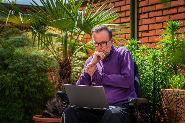 A male disabled person with dystrophy sits in his atrium in a wheelchair behind his brick wall, among the greenery, communicates with advice via a notebook on his knees and with a telephone, observes the surroundings and greenery stock photo