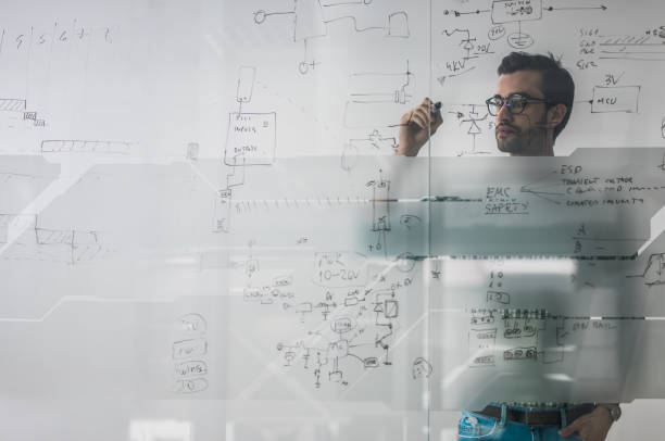 Male engineer working on new ideas and writing diagram on glass wall. Young engineer drawing a diagram on a glass in the office. The view is through glass. complexity stock pictures, royalty-free photos & images