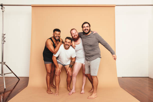 Male friends laughing together in a studio Male friends laughing cheerfully in a studio. Four body positive men having fun while standing together. Group of self-confident young men feeling comfortable in their natural bodies. body positive stock pictures, royalty-free photos & images
