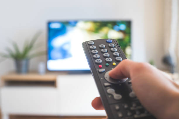 Male hand is holding TV remote control, streaming on a smart TV. Holding a TV remote control in the hand, foreground, tv in the blurry background. Streaming. video on demand stock pictures, royalty-free photos & images