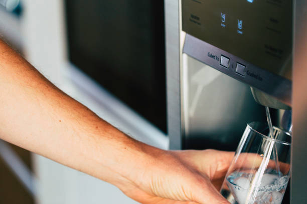 Male hand is pouring cold water and ice cubes from dispenser of home fridge. stock photo