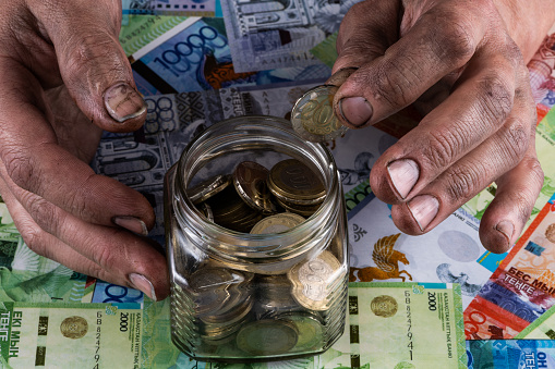 Male hands of a working man pull out a 200 Kazakhstan tenge coin from a glass jar with coins against the background of Kazakhstan banknotes