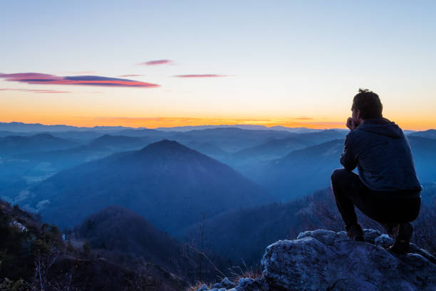 Male hiker crouching on top of the hill watching twilight landscape after sunset stock photo
