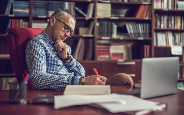 Male manager taking notes in home office. Mid adult manager sitting at home office and writing something in a notebook. professor stock pictures, royalty-free photos & images