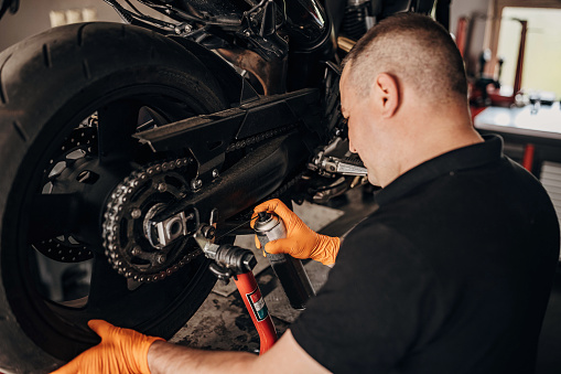 One man, male motorcycle mechanic repairing motorcycle in his workshop. Spraying oil on chain.
