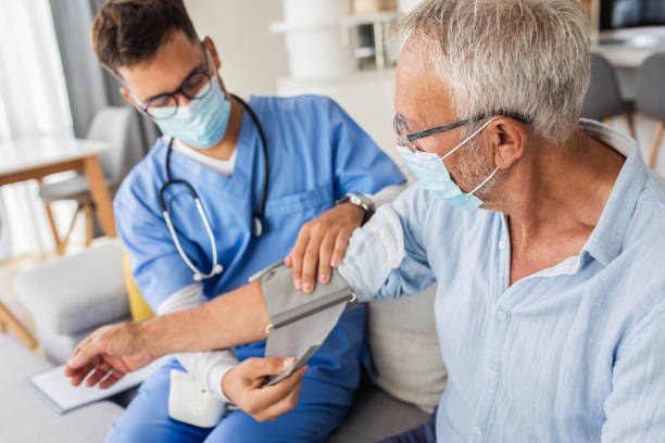 Male nurse measures blood pressure to senior man with mask while being in a home visit. Male nurse measures blood pressure to senior man with mask while being in a home visit. blood pressure gauge stock pictures, royalty-free photos & images
