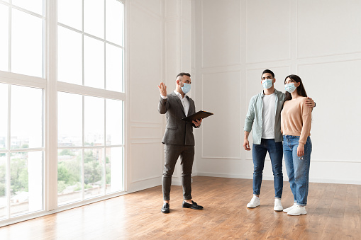 Meeting With Realtor During Pandemic. Professional Real Estate Agent In Suit And Face Mask Showing Modern House With Floor-to-Ceiling Windows To Beautiful Young Couple Purchasing Or Renting New Home
