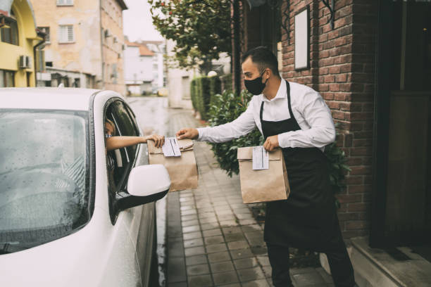 Male restaurant employee delivers packed food to a female driver outside a restaurant. Waiter wearing protective face mask is giving disposable package with food  to pretty female driver outside a restaurant. curbsidepickup stock pictures, royalty-free photos & images