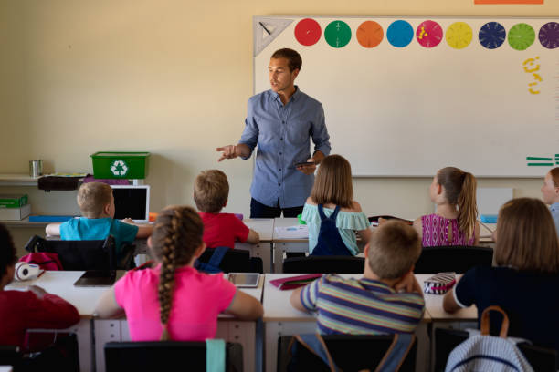 Male school teacher standing in an elementary school classroom with a group of school children Front view of a Caucasian male school teacher standing and addressing a diverse group of school children sitting at desks during a lesson in an elementary school classroom in front of stock pictures, royalty-free photos & images