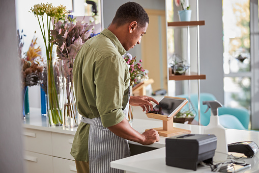 Young handsome man in apron is standing at counter with tablet and POS terminal while selling bouquets