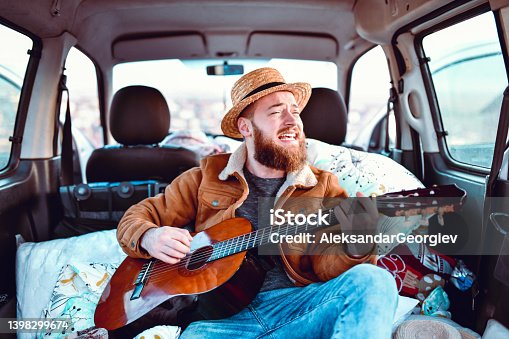 istock Male Smiling And Singing While Playing Guitar In Minivan Trunk 1398299674