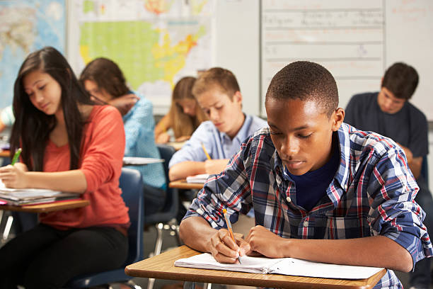 Male student in classroom writing in notebook Teenage African American male wearing a plaid button-up shirt sitting at a desk in class. He is writing in a notebook on his desk using a pencil.  Around him are his classmates, also writing on their papers.  They are all concentrating on their own work. high school student stock pictures, royalty-free photos & images