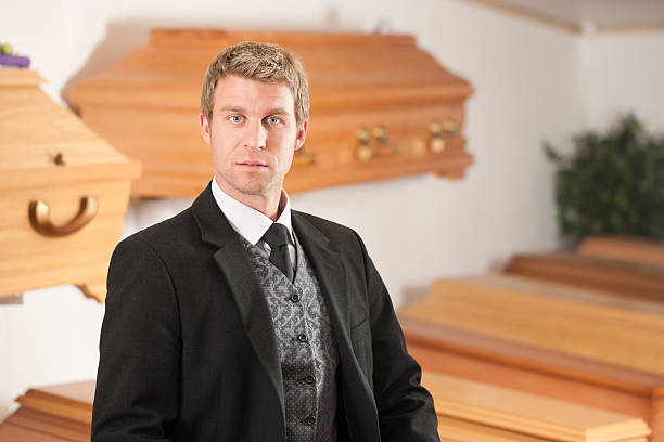 Male suited undertaker with coffins in background Undertaker in his store with a display of coffins undertaker stock pictures, royalty-free photos & images