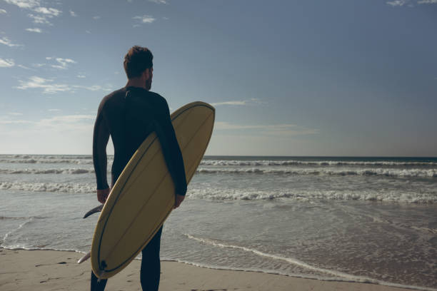 Male surfer with a surfboard standing on a beach Rear view of young Caucasian male surfer in wet suit with a surfboard standing face to the ocean on a beach on sunny day. He is holding his surfboard Smart Surf Suit stock pictures, royalty-free photos & images
