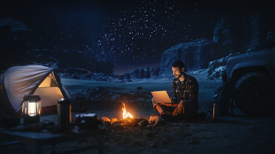 Male Traveler Sitting by Campfire Uses Laptop Computer while Tent Camping in the Canyon. Man doing Digital Remote Work, e-business, e-shopping, ecommerce through Internet while being on Vacation Trip