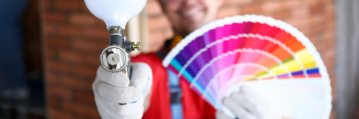 Close-up of smiling construction site worker holding colourful palette with samples and empty professional airbrush gun. Renovation and interior design concept
