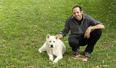 A man in his thirties kneeling next to a large white akita that is laying in the green grass.