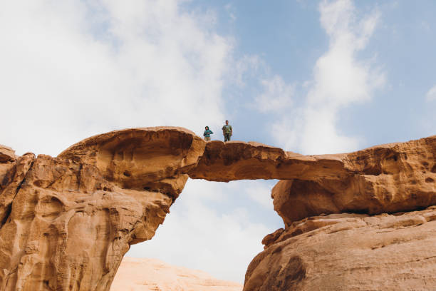 Man and woman traveler staying at the natural arch enjoying a view of Wadi Rum desert Female and male explorers contemplating the arid landscape walking above the arch in Jordan hot middle eastern women stock pictures, royalty-free photos & images