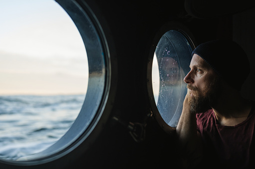 Man at the porthole window of a vessel sailing the sea, watching out in a pensive mood.