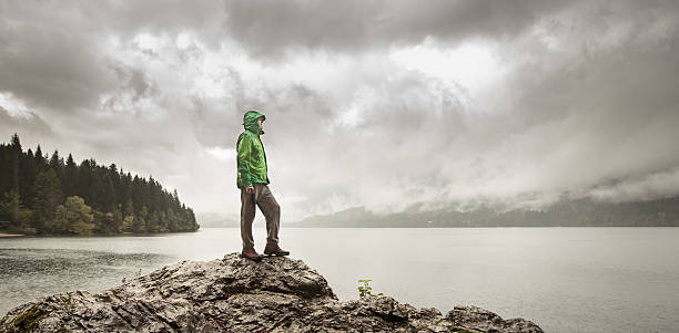 Man beside a mountain lake in rain Man standing on a rock beside a dramatic mountain lake after a hike in the rainy, gloomy day. Active lifestyle, outdoor activities, moods and emotions concept. storm watching stock pictures, royalty-free photos & images