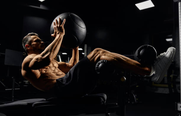 Man building core muscles with ball. Side view of muscular caucasian man training abs with ball on bench in gym. Handsome young sportsman building core muscles, doing crunches in dark atmosphere. Concept of sport, fitness. male bodybuilders stock pictures, royalty-free photos & images