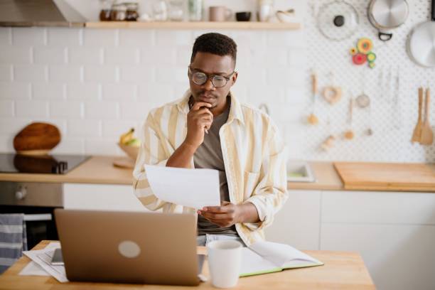 Man calculates personal income by using laptop and online banking on smartphone African man working from home, using laptop, paying utility bills. Young man doing taxes at home. personal loan stock pictures, royalty-free photos & images