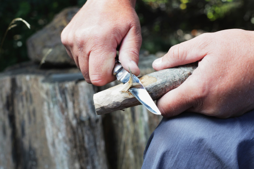 A man uses a bowie knife with a sharp blade to carve a wooden stick.