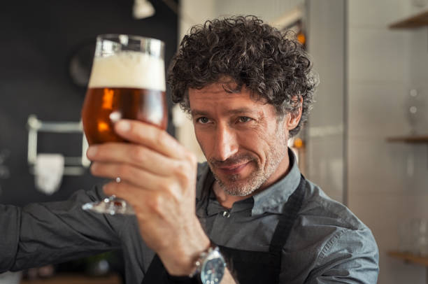 Man checking beer quality Mature man examining the quality of craft beer at brewery. Inspector working at alcohol manufacturing factory checking beer. Man in distillery checking quality control of draught beer. artisanal food and drink stock pictures, royalty-free photos & images