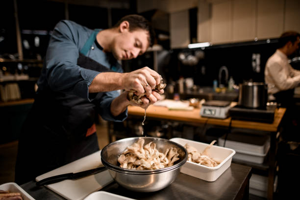 man chef holds handful of oyster mushrooms over bowl on kitchen man chef holds handful of oyster mushrooms over bowl on kitchen. Vegan dishes with oyster mushrooms oyster mushroom stock pictures, royalty-free photos & images