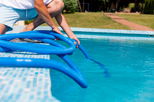 Mature man in flip flops cleaning the swimming pool with a vacuum cleaner.  Man working as a cleaner of the swimming pool, he standing with special equipment for cleaning at poolside and working
