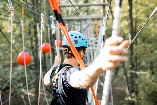 Well-equipped man having an active recreation, climbing in a rope park with obstacles in the forest
