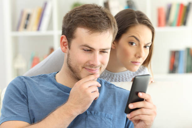 Man dating on line and girlfriend spying Cheater man dating on line with a smart phone and girlfriend is spying sitting on a sofa at home envy stock pictures, royalty-free photos & images