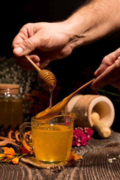 Man hand holding wooden honey dipper, honey spoon on top of glass of tea and dripping honey in hot tea stock photo