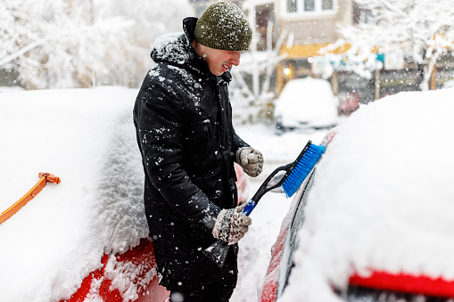 Man is Cleaning Snow from a Car with a Brush. Man Outdoors is Cleaning Snow on his Car Windshield.