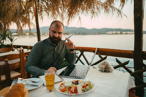 Young man sitting at the restaurant, using a laptop, and having an online class during summer vacation at the seaside
