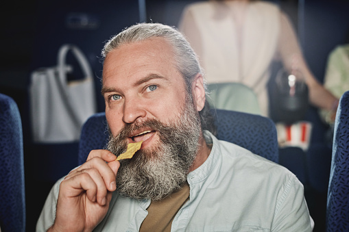 Close-up of mature Caucasian man with beard on face enjoying having snack and watching film at cinema