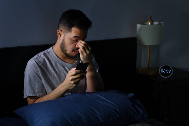 Man having sore and tired eyes when using smartphone while lying in bed at night stock photo
